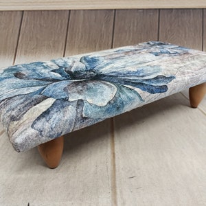 May include: A small, rectangular footstool with a floral design in shades of blue and gray. The footstool has four wooden legs.