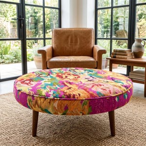 Puede incluir: Un otomán redondo con una tapicería floral multicolor en tonos magenta, naranja, amarillo y verde. La pieza se apoya sobre cuatro patas cónicas de madera oscura y presenta una silueta circular de perfil bajo.