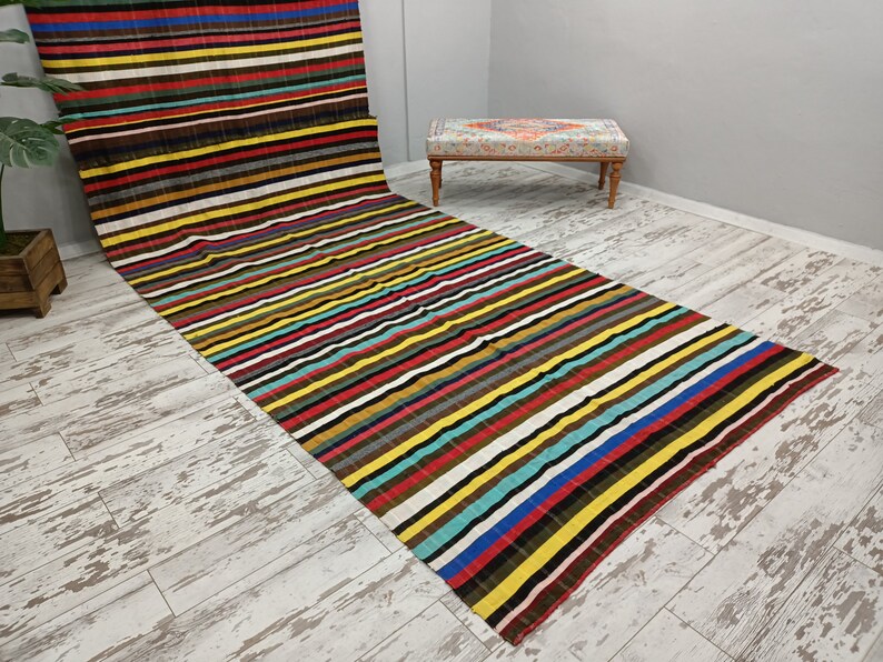 May include: A colorful striped rug with a variety of colors, including red, yellow, blue, green, and brown. The rug is laid out on a whitewashed wood floor.