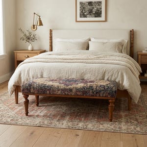May include: A bedroom with a bed featuring a cream headboard and bedding. A patterned bench with wooden legs is at the foot of the bed. A rug with a faded red and cream pattern is on the floor.