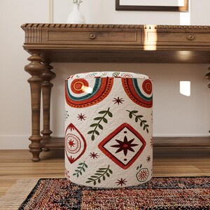 May include: A cylindrical ottoman featuring a cream-coloured fabric base with a repeating pattern of red, teal, and dark green geometric shapes, including diamonds, concentric semicircles, starbursts, and leafy sprigs across the entire surface.