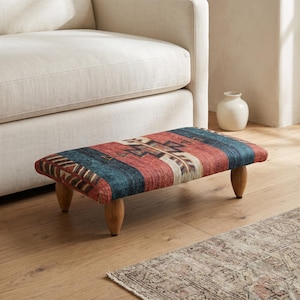 May include: A rectangular footstool featuring a woven fabric top with a geometric pattern in shades of red, blue, and cream. The footstool is supported by four short, rounded wooden legs with a natural light brown finish.