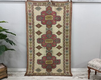 Puede incluir: Una alfombra turca vintage con fondo crema y un patrón geométrico en tonos rojos, azules y amarillos. La alfombra tiene un borde de flecos y está colgada en una pared blanca.