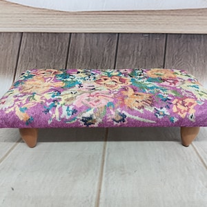 Puede incluir: Un reposapiés con estampado floral y patas de madera. El reposapiés es morado con una variedad de flores de colores.