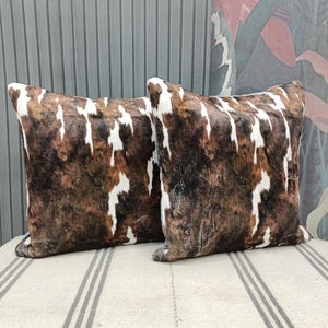 May include: Two brown and white cowhide patterned throw pillows with a white and gray striped background.