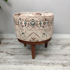 May include: A round ottoman with a beige fabric cover featuring a colourful floral pattern. The ottoman has a wooden base with three legs.
