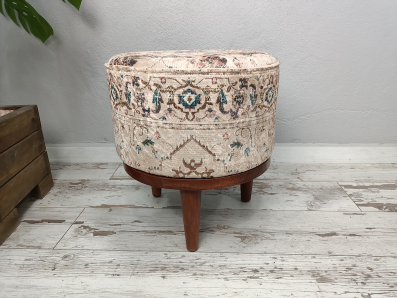 May include: A round ottoman with a beige fabric cover featuring a colourful floral pattern. The ottoman has a wooden base with three legs.