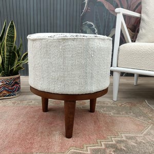 Puede incluir: Un otomán redondo tapizado en blanco con base y patas de madera marrón oscuro. El otomán está colocado sobre una alfombra estampada en rosa y gris. Una planta en maceta y una silla blanca están en el fondo.