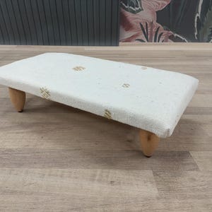 May include: A rectangular, off-white footstool with a textured fabric top and four short, rounded wooden legs. The fabric is decorated with small, gold-colored floral patterns. The stool is designed for comfort and style.
