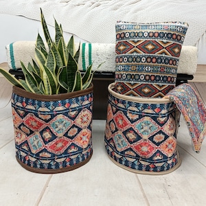 May include: Two cylindrical storage baskets with a blue, red and white geometric pattern. The baskets are made of fabric and have a brown leather trim. A green snake plant is in one of the baskets. A cushion with a similar pattern is on top of the baskets.