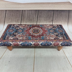 May include: A small, rectangular ottoman with a colorful, patterned fabric top. The fabric features a central star design with blue, red, and cream colors. The ottoman has wooden legs.