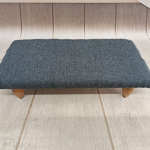 Puede incluir: Un reposapiés tapizado en gris con patas de madera. El reposapiés está sobre un suelo de madera clara.