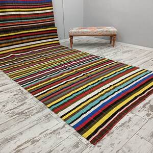 May include: A colorful striped rug with a variety of colors, including red, yellow, blue, green, and brown. The rug is laid out on a light wood floor.