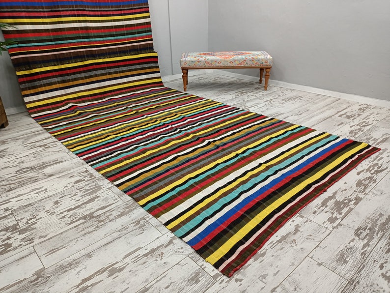 May include: A colorful striped rug with a variety of colors, including red, yellow, blue, green, and brown. The rug is laid out on a light wood floor.