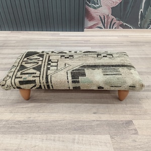 May include: A rectangular footstool with a patterned, woven top in shades of black, beige, and green. The footstool is supported by four short, rounded wooden legs. The design features geometric shapes and lines.