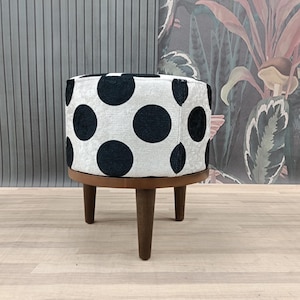 Puede incluir: Un pequeño otomán redondo con una funda de tela blanca con grandes lunares negros. El otomán tiene una base de madera marrón oscuro y tres patas cónicas. El diseño es una mezcla de estilos modernos y retro.
