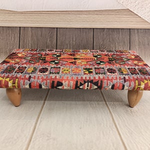 May include: A red, yellow, and blue patterned ottoman with wooden legs. The ottoman is made of fabric and has a traditional design.