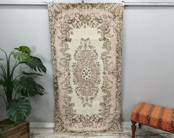May include: A rectangular, vintage-style rug with a central floral medallion design. The rug features a cream-colored field with a pink border and brown floral accents. The rug is displayed against a gray wall.