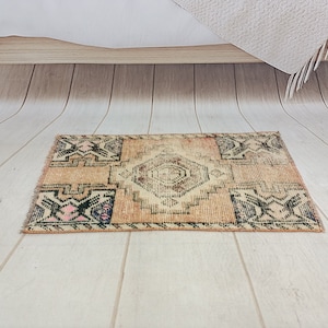 May include: A small, rectangular rug with a beige background and a black and pink geometric pattern. The rug is on a light wood floor.