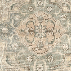 May include: A vintage rug with a beige and green color scheme. The rug features a central medallion with a floral design. The rug has a distressed look, with some areas showing wear and tear.