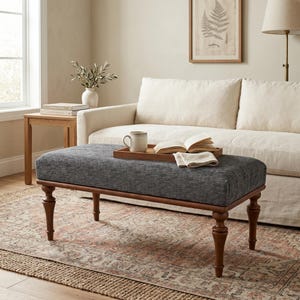 May include: A gray upholstered ottoman with a dark wood frame and turned legs. The ottoman is rectangular and sits on a patterned rug. A wooden tray with a mug and book rests on top.