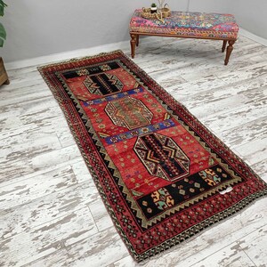 May include: A red, black, and blue patterned runner rug with a geometric design. The rug has a fringe border and is laid out on a whitewashed wood floor.