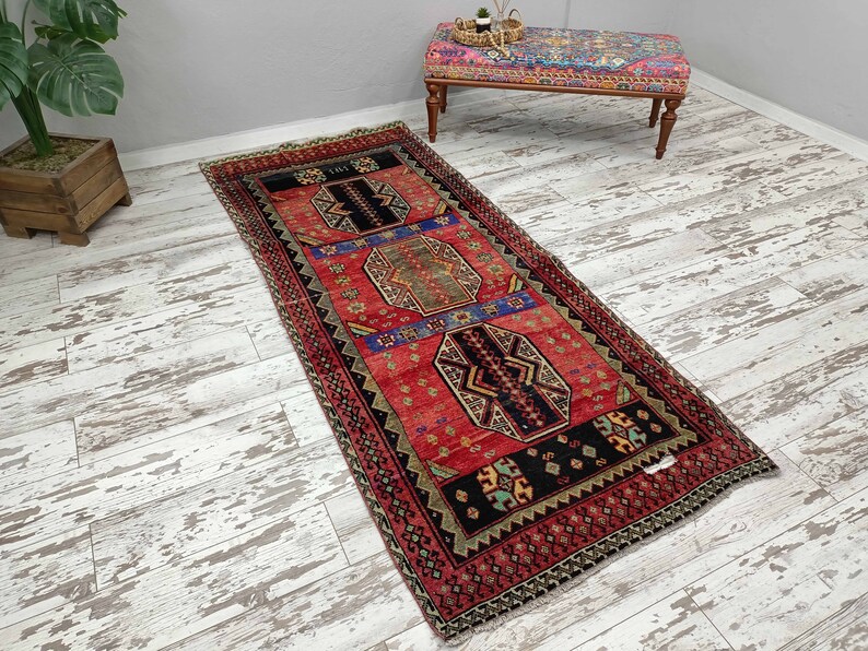 May include: A red, black, and blue patterned runner rug with a geometric design. The rug has a fringe border and is laid out on a whitewashed wood floor.