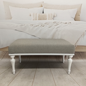 May include: A white upholstered bench with turned legs and a grey fabric top. The bench is in front of a bed with white bedding and a throw blanket.