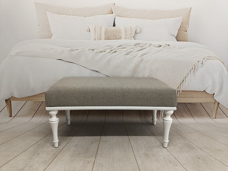 May include: A white upholstered bench with turned legs and a grey fabric top. The bench is in front of a bed with white bedding and a throw blanket.