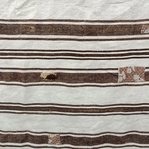 Puede incluir: Primer plano de un textil a rayas blancas y marrones. El textil tiene rayas marrones horizontales de diferentes anchos, alternando con rayas blancas m&aacute;s anchas. Parches de tela estampada est&aacute;n cosidos en el textil. Parece ser una alfombra o una manta.