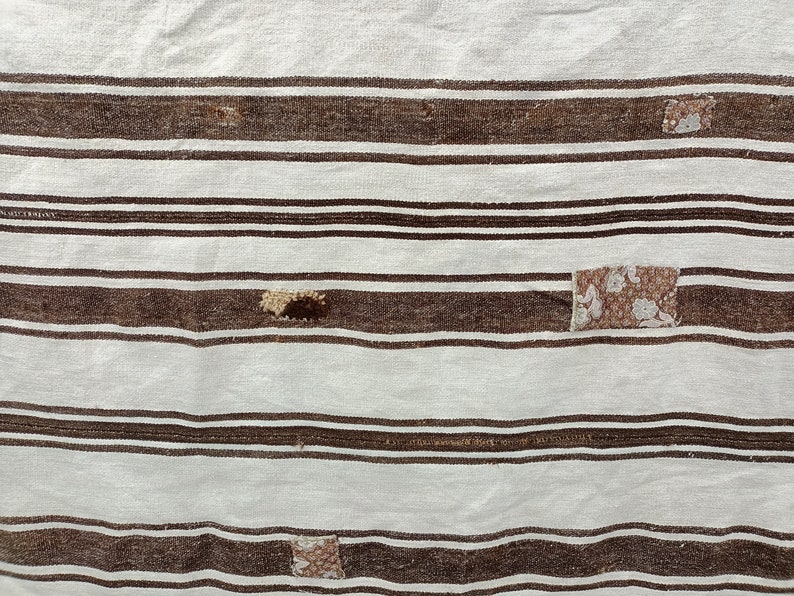 Puede incluir: Primer plano de un textil a rayas blancas y marrones. El textil tiene rayas marrones horizontales de diferentes anchos, alternando con rayas blancas m&aacute;s anchas. Parches de tela estampada est&aacute;n cosidos en el textil. Parece ser una alfombra o una manta.