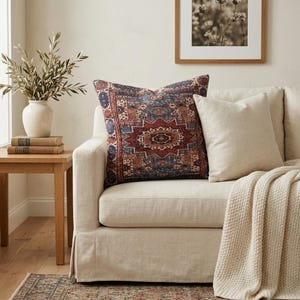 a cozy living room with a beige sofa adorned with a colorful patterned pillow and a white throw blanket. A wooden side table holds a vase with greenery and a stack of books. A framed picture hangs on the wall above the sofa, adding a personal touch to the space.