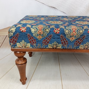 May include: A blue and gold patterned ottoman with wooden legs. The ottoman has a floral design with red accents.