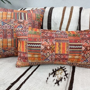 Op de afbeelding: Twee decoratieve sierkussens met een patchwork-ontwerp. De kussens hebben een mix van oranje, rode en gele tinten met geometrische en tribale patronen. De kussens zijn rechthoekig en vierkant en liggen op een wit en zwart gestreept oppervlak.