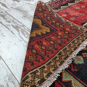 May include: A red, black, and gold patterned rug with a fringe border. The rug is folded in half and the fringe is visible on the edge.