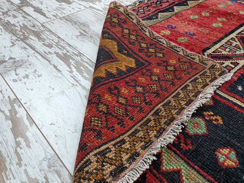 May include: A red, black, and gold patterned rug with a fringe border. The rug is folded in half and the fringe is visible on the edge.