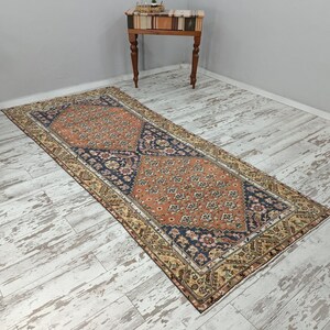 May include: A vintage runner rug with a traditional Persian design. The rug has a cream, orange, and blue color scheme with intricate floral patterns. The rug is laid out on a whitewashed wood floor.