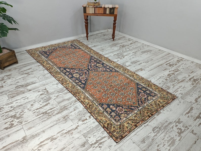 May include: A vintage runner rug with a traditional Persian design. The rug has a cream, orange, and blue color scheme with intricate floral patterns. The rug is laid out on a whitewashed wood floor.