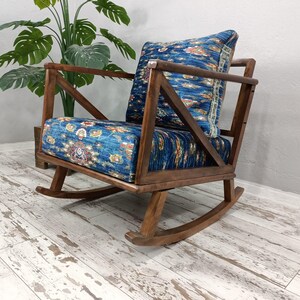 A wooden rocking chair with a blue and white patterned cushion sits on a white wooden floor, with a potted plant in the background.