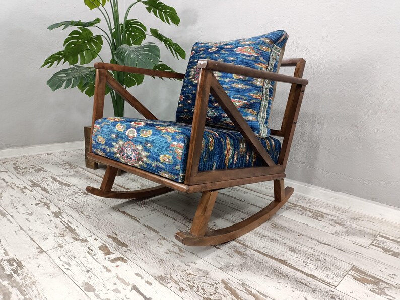 A wooden rocking chair with a blue and white patterned cushion sits on a white wooden floor, with a potted plant in the background.