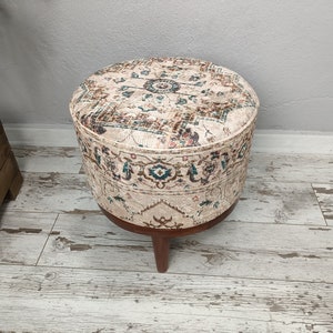 May include: A round, cream-coloured ottoman with a brown wooden base and intricate floral pattern. The ottoman is sitting on a light wood floor.