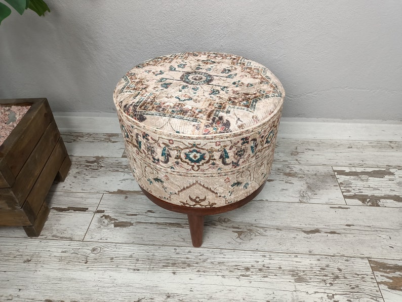 May include: A round, cream-coloured ottoman with a brown wooden base and intricate floral pattern. The ottoman is sitting on a light wood floor.