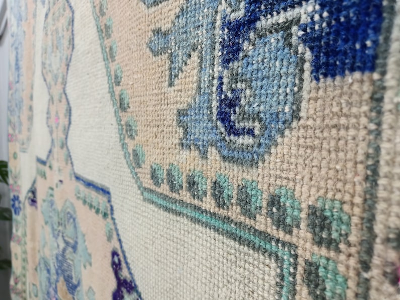 K&ouml;nnte beinhalten: Nahaufnahme eines cremefarbenen Teppichs mit einem blauen, gr&uuml;nen und rosafarbenen Blumenmuster. Der Teppich hat eine gewebte Textur und besteht aus weichen Fasern.