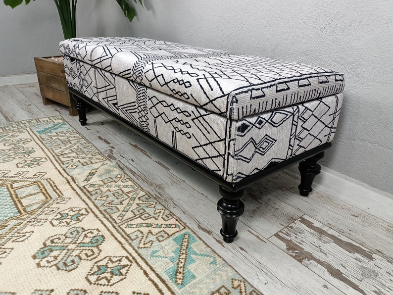 May include: A black and white patterned storage bench with black turned legs. The bench is sitting on a light wood floor with a patterned rug in front of it.