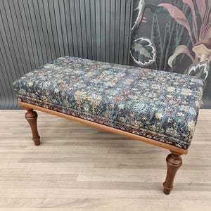 Könnte beinhalten: Ein rechteckiger Hocker mit einem gemusterten Stoffbezug in Blau-, Rot- und Goldtönen. Der Holzrahmen hat eine hellbraune Oberfläche und die Beine sind dunkelbraun. Der Hocker ist ein dekoratives Möbelstück.