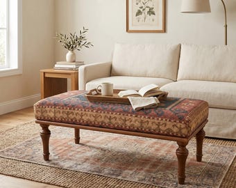 May include: A rectangular ottoman with a patterned fabric top in shades of red, blue, and tan, resting on four dark wooden legs. A wooden tray with a mug and book sits on top. The ottoman is placed on a rug in a living room setting.