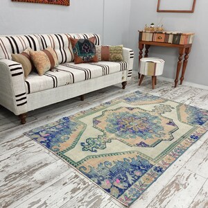 K&ouml;nnte beinhalten: Ein Vintage-Teppich mit hellbeigefarbenem Hintergrund und einem blauen und rosafarbenen Blumenmuster. Der Teppich liegt auf einem wei&szlig; get&uuml;nchten Holzboden in einem Wohnzimmer mit einem wei&szlig;en Sofa und einem kleinen Tisch mit einem Hocker.