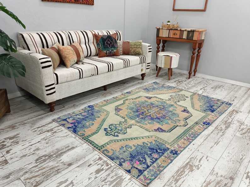 K&ouml;nnte beinhalten: Ein Vintage-Teppich mit hellbeigefarbenem Hintergrund und einem blauen und rosafarbenen Blumenmuster. Der Teppich liegt auf einem wei&szlig; get&uuml;nchten Holzboden in einem Wohnzimmer mit einem wei&szlig;en Sofa und einem kleinen Tisch mit einem Hocker.
