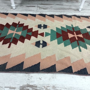 K&ouml;nnte beinhalten: Ein rechteckiger Teppich mit geometrischem Muster. Der Teppich hat einen cremefarbenen Hintergrund mit einem zentralen Design in T&uuml;rkis, Burgunderrot und Marineblau. Der Rand und die Zickzackkanten sind in Rosa- und Marineblaut&ouml;nen gehalten.