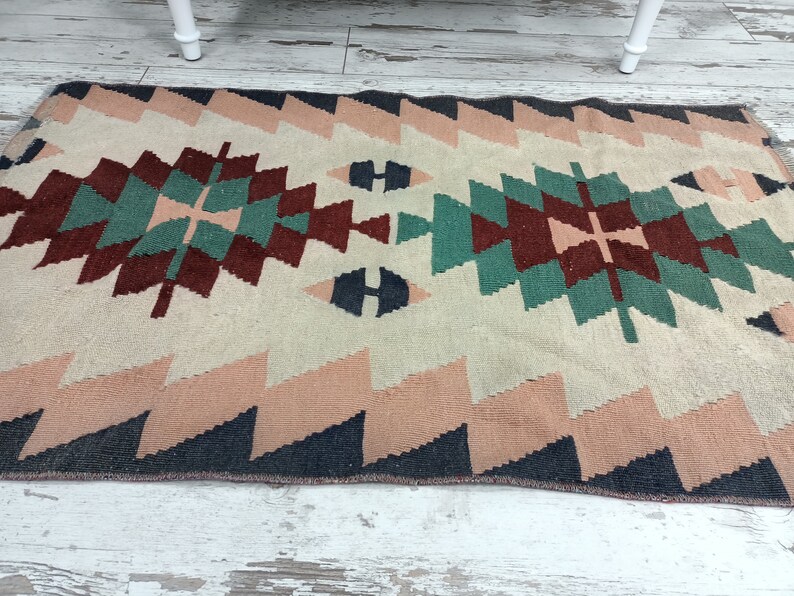 K&ouml;nnte beinhalten: Ein rechteckiger Teppich mit geometrischem Muster. Der Teppich hat einen cremefarbenen Hintergrund mit einem zentralen Design in T&uuml;rkis, Burgunderrot und Marineblau. Der Rand und die Zickzackkanten sind in Rosa- und Marineblaut&ouml;nen gehalten.